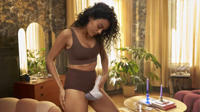 Mujer con ropa interior marrón usa un aparato blanco. Fondo con cortinas doradas, plantas y una mesa con velas.