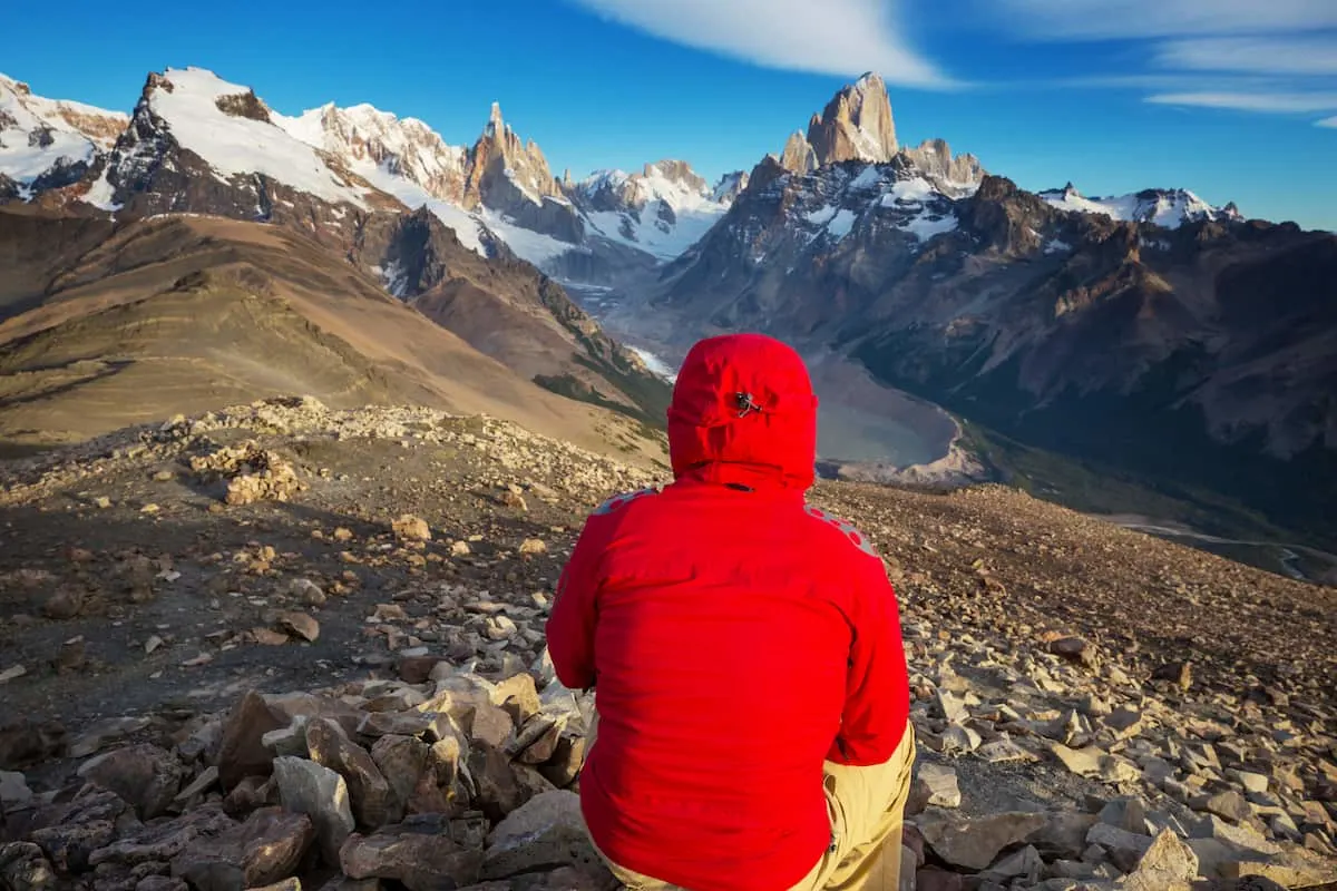 Persona con chaqueta roja de espaldas sentada sobre un terreno rocoso, contemplando una cordillera de montañas nevadas bajo un cielo azul.
