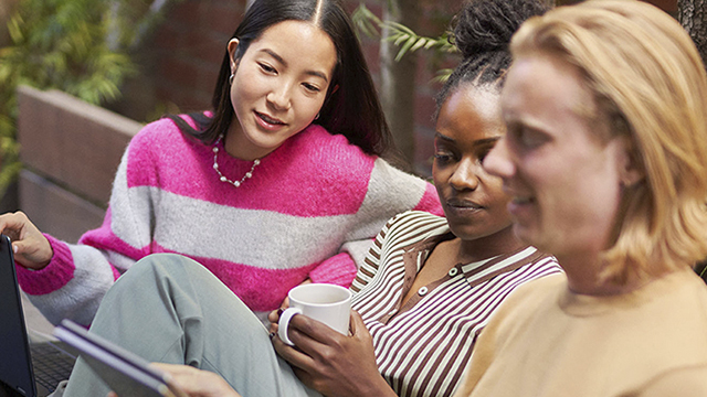 Tres personas están sentadas al aire libre. Una sostiene una taza, otra un portátil y la tercera está cerca.