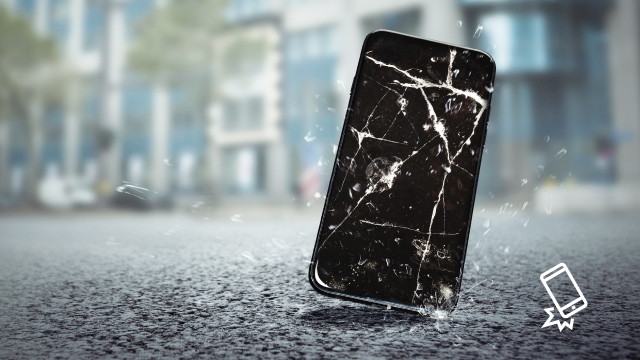 Un smartphone negro con la pantalla rota está apoyado sobre una superficie gris, con edificios borrosos al fondo.