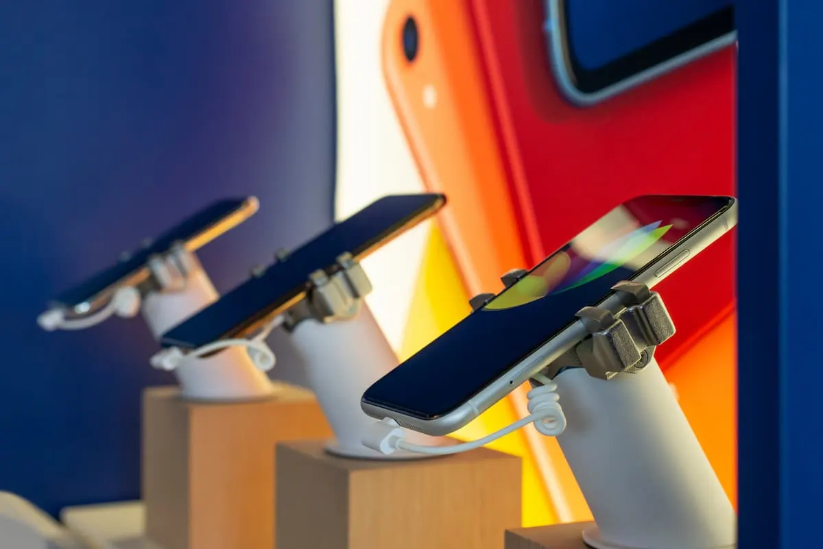 Tres smartphones en exposición sobre soportes de seguridad blancos, alineados en una mesa de tienda de electrónica, con una pantalla colorida y desenfocada al fondo.