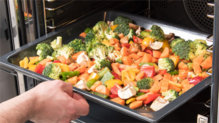 Detalle de una mano introduciendo una bandeja de horno con hortalizas y vegetales
