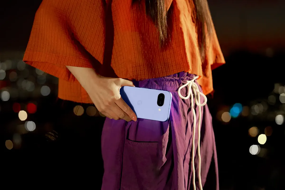 Una persona guarda un teléfono lila en el bolsillo de un pantalón morado. Una camisa naranja completa el atuendo. Luces borrosas al fondo.