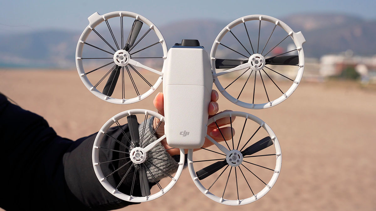 Una persona sostiene un dron blanco con cuatro rotores protegidos por aros, sobre un fondo difuminado de playa y colinas.