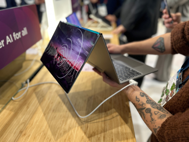 Un portátil abierto con un diseño abstracto en la pantalla, conectado a un cable blanco. Una persona lo sostiene.