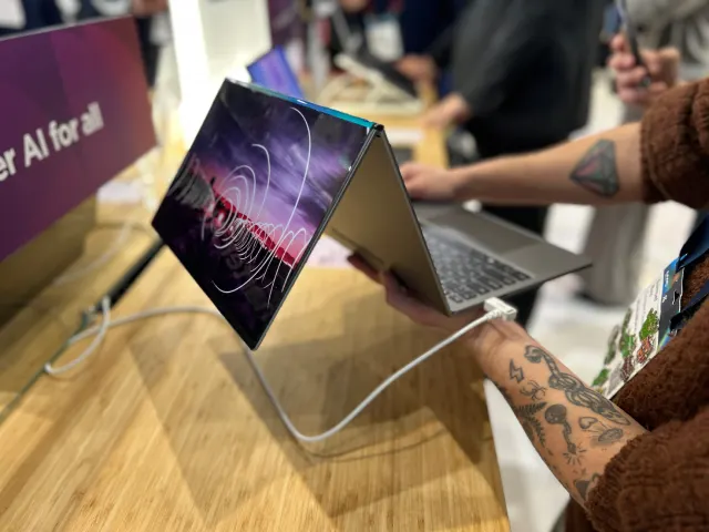 Un portátil abierto con un diseño abstracto en la pantalla, conectado a un cable blanco. Una persona lo sostiene.