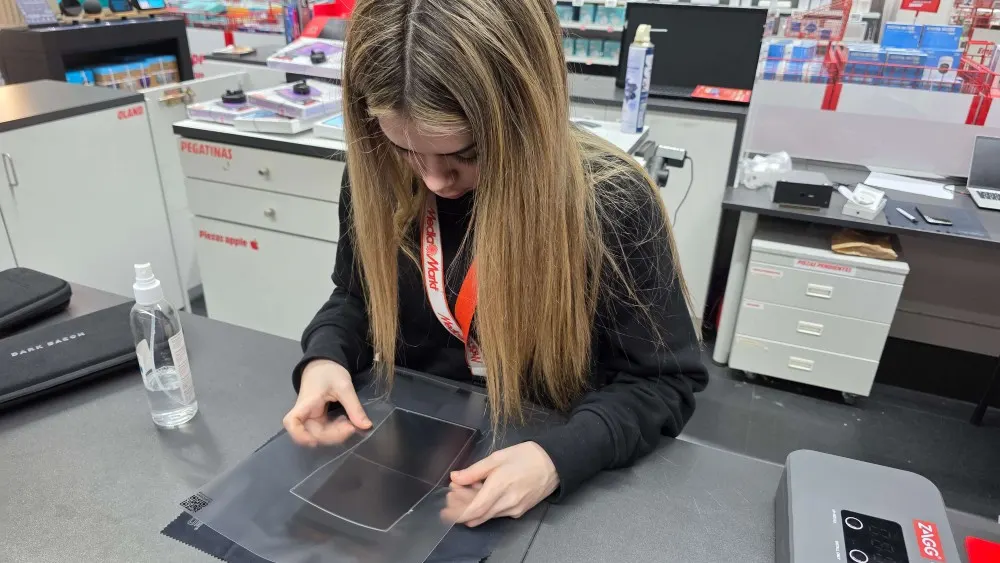 Especialista del Service Point de MediaMarkt instalando cuidadosamente un protector de pantalla sobre una consola en el mostrador, con herramientas y productos de limpieza alrededor.