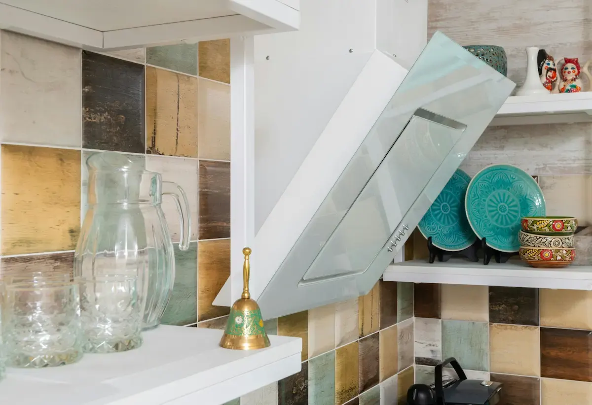 Estantes con jarra de cristal, vasos y campana extractora blanca sobre azulejos multicolores. Platos decorativos en estante superior.