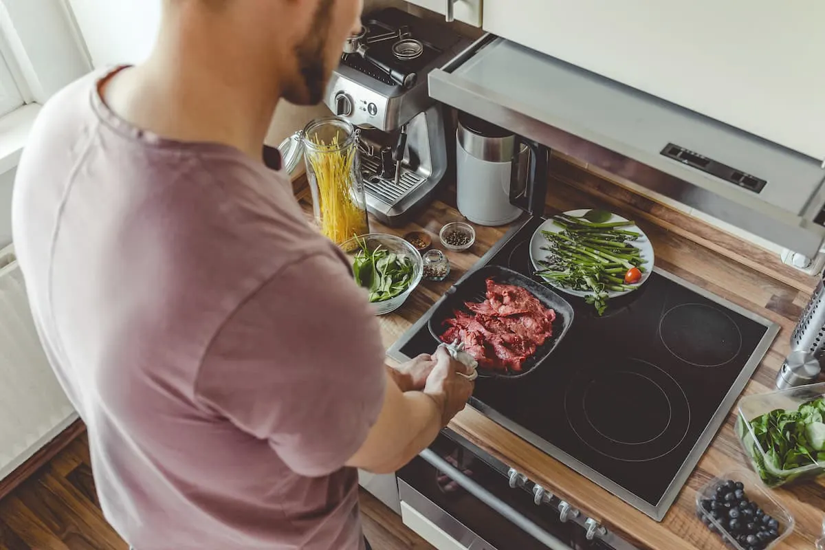 Hombre cocinando carne en sartén sobre placa de inducción en cocina moderna, rodeado de verduras frescas y utensilios.