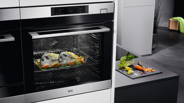 Horno AEG empotrado con comida en una bandeja y verduras cortadas en una tabla negra. Se aprecia una nevera blanca al lado.