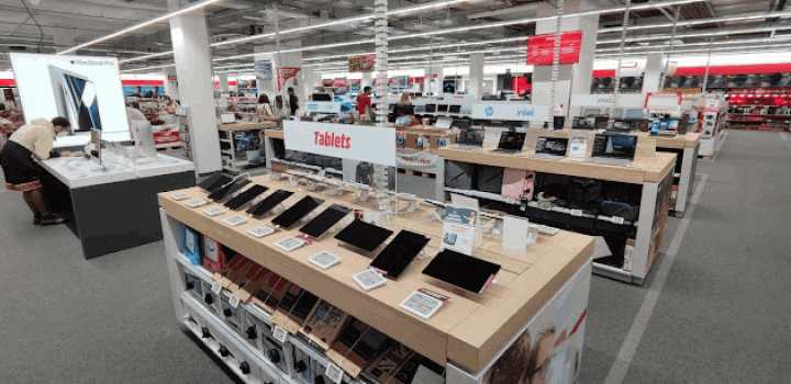 Interior de tienda con mostradores de tablets y computadoras, personal y clientes presentes.