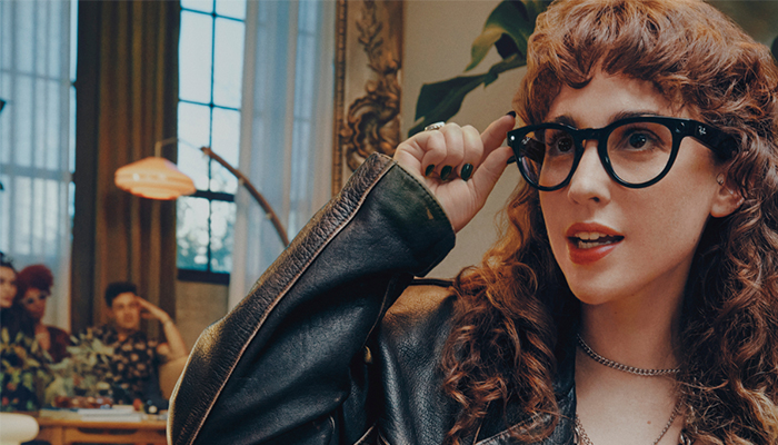 Una persona con gafas y chaqueta de cuero, con otras personas y una lámpara visibles al fondo.