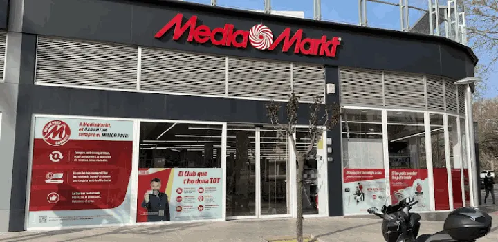 Fachada de tienda Media Markt con rótulo rojo. Ventanas con carteles promocionales y persianas metálicas. Una moto aparcada delante.