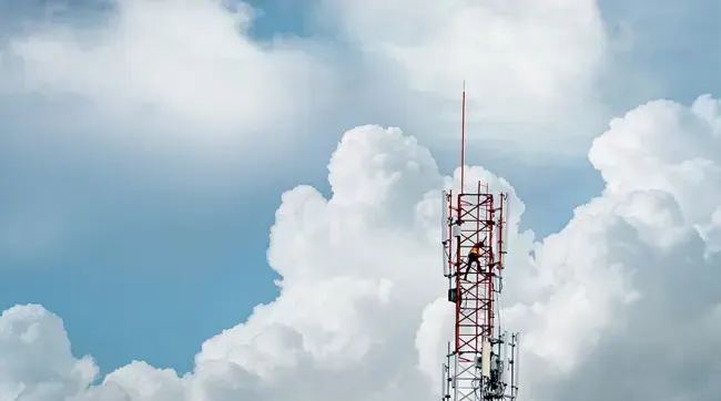 Torre de telecomunicaciones roja contra un cielo azul con nubes blancas y esponjosas. Una persona está trabajando en la torre.