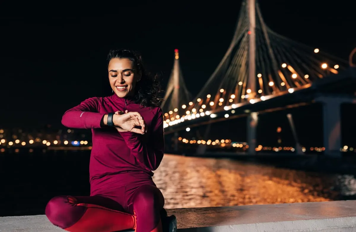 Mujer deportista revisando su smartwatch sentada al aire libre de noche con un puente iluminado de fondo.