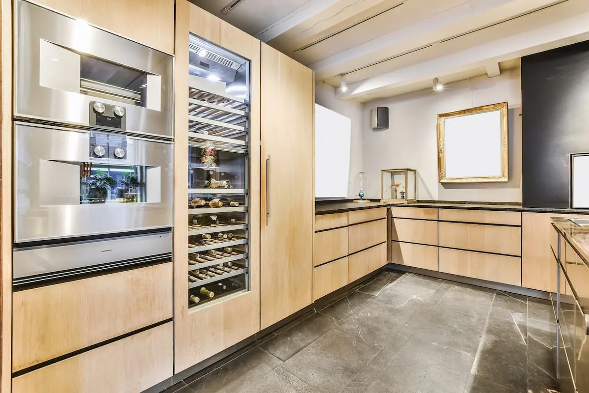 Cocina de madera clara con vinoteca integrada, horno de acero inoxidable y suelo de piedra oscura.