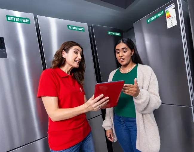 Dos mujeres en una tienda de electrodomésticos, mirando una tableta roja frente a refrigeradores de acero inoxidable.