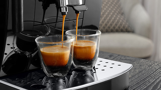 Máquina de café negra sirviendo café en dos vasos de vidrio sobre una bandeja metálica con agujeros.