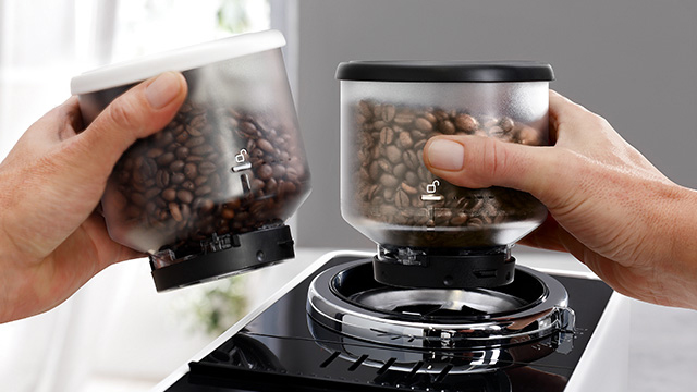 Dos manos sostienen recipientes transparentes llenos de granos de café sobre una máquina negra.