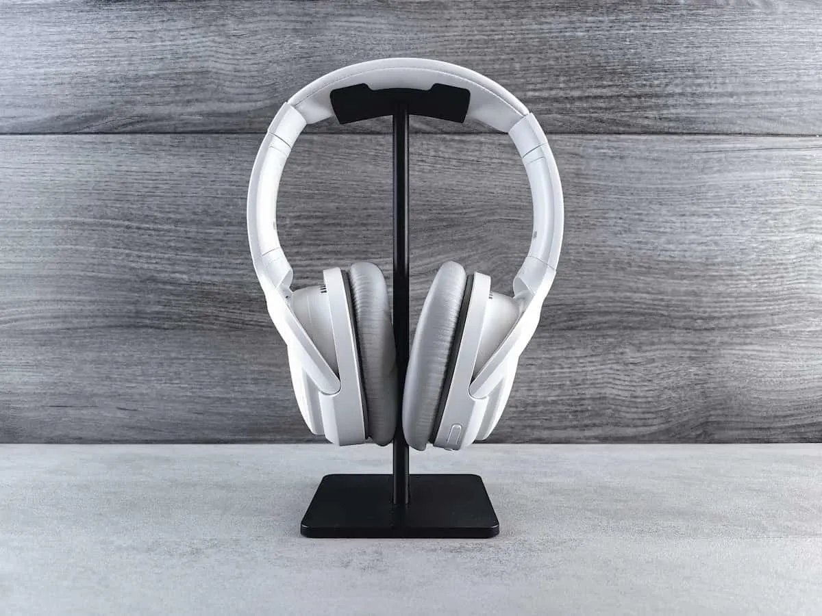 Auriculares blancos de diadema colocados sobre un soporte negro vertical, con fondo de superficie gris y pared con textura de madera gris.