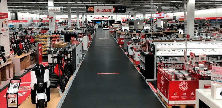 Vista interior de una tienda grande con pasillos llenos de estanterías con productos variados. El suelo es gris oscuro.