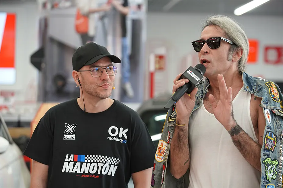 Dos hombres en un evento. Uno con gorra negra y camiseta con logos. El otro con gafas de sol y chaqueta vaquera con parches, sosteniendo un micrófono.