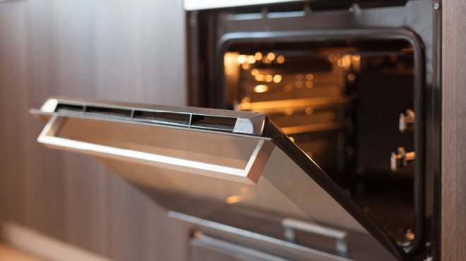 Horno abierto con luz interior, puerta abatible gris y rejillas visibles. El mueble circundante es de tono madera.