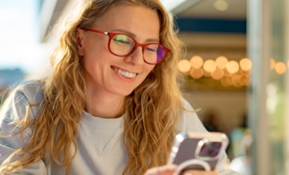 Mujer sonriendo con gafas rojas usando un móvil. Texto superpuesto: "Detector de fraudes por SMS" y logo de McAfee Smart KI. Fondo urbano.