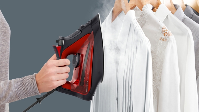 Una persona plancha una prenda roja con una plancha azul. Vapor visible. Un reloj rojo está sobre una encimera blanca al fondo.