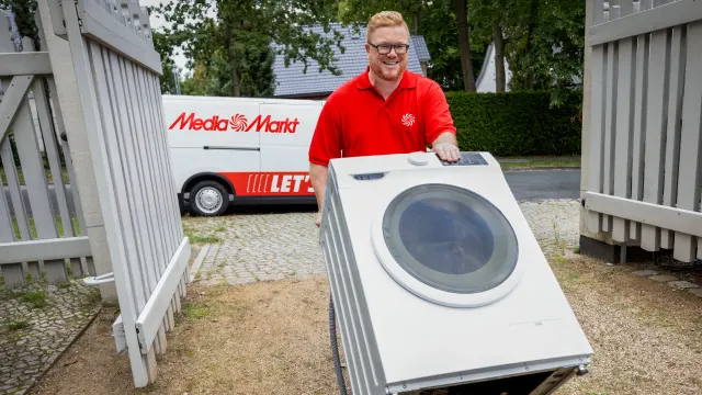Empleado de MediaMarkt con uniforme rojo presenta una lavadora blanca frente a un vehículo de entrega de la empresa en una vivienda.