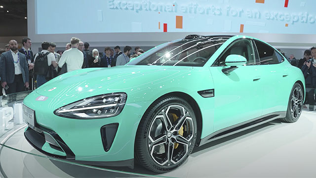 Coche turquesa en exhibición, rodeado de personas y con un fondo de pantalla azul con texto y formas geométricas.