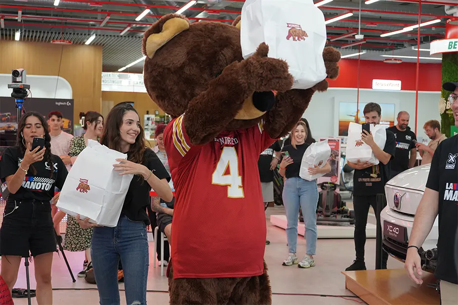 Un oso de peluche con camiseta roja y número 4 levanta bolsas blancas ante un grupo de personas en un espacio interior.