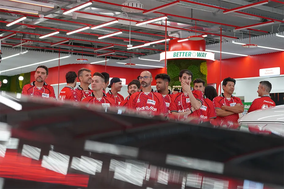 Un grupo de personas con camisetas rojas se encuentra en un lugar con techo blanco y detalles rojos. Se ve un letrero que dice "Better Way".