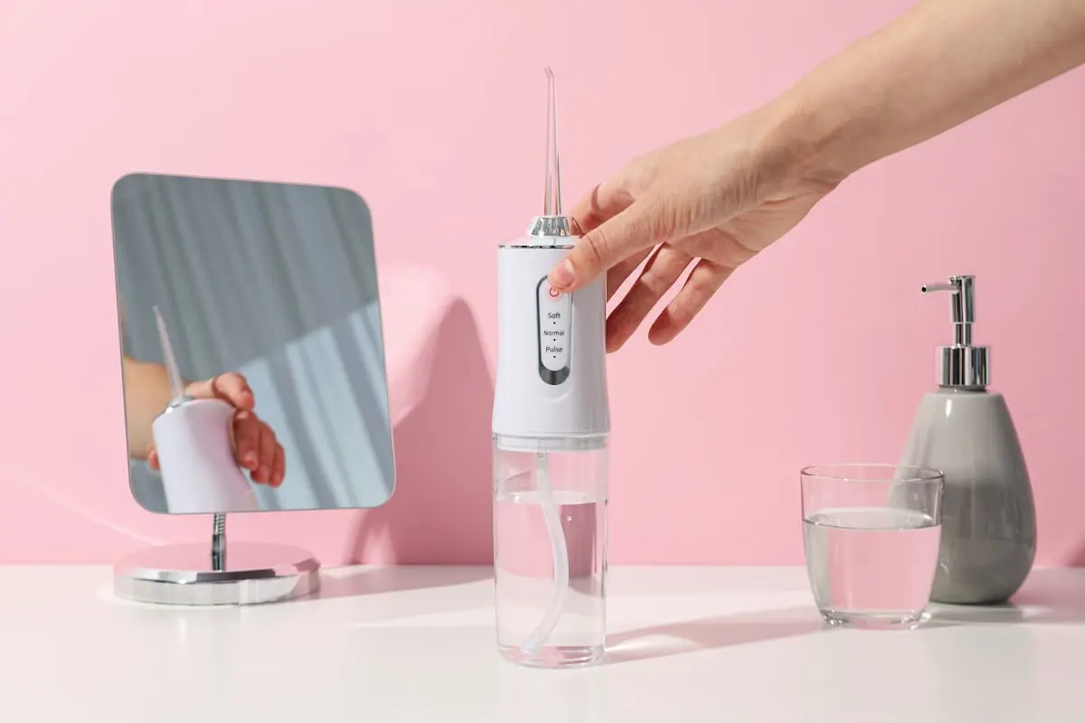 Mano sosteniendo un irrigador dental blanco sobre una mesa con fondo rosa.