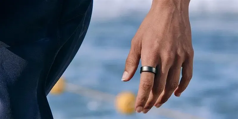 Mano con anillo metálico en el dedo, sobre un fondo azul difuminado con elementos amarillos esféricos fuera de foco.