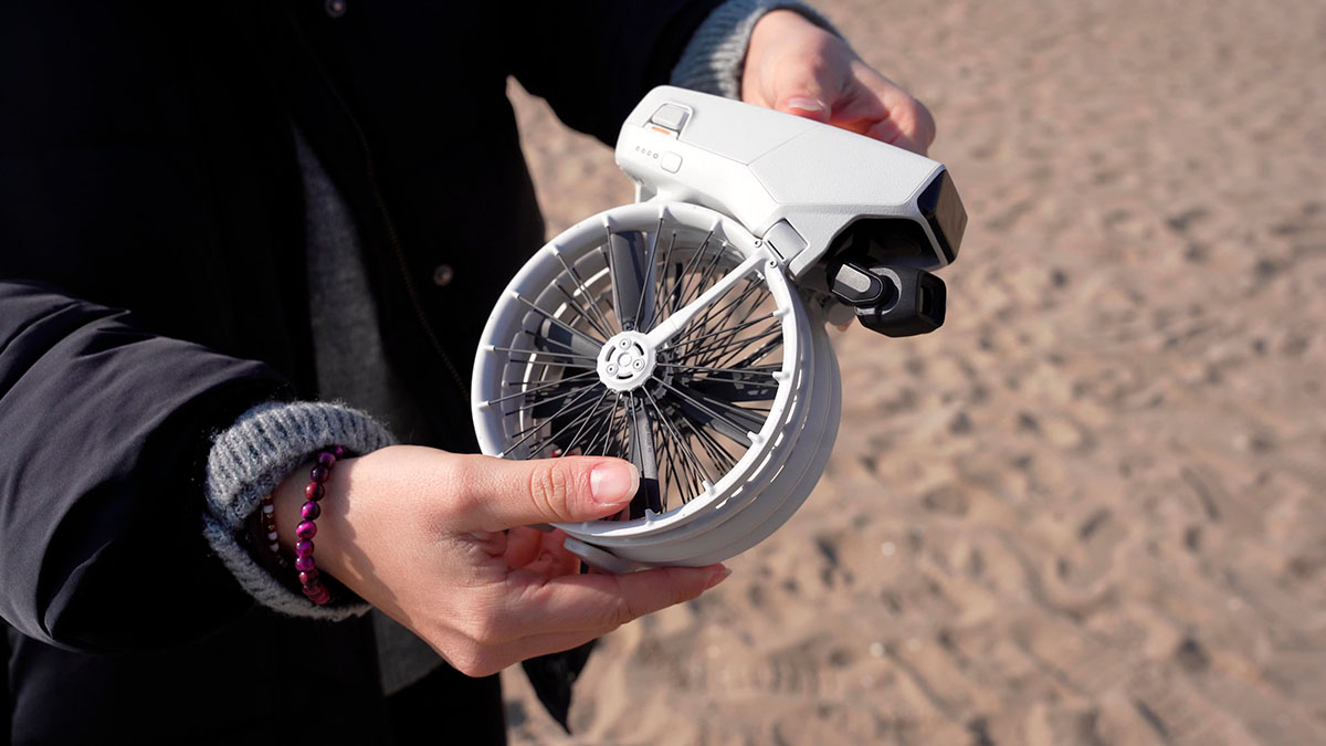 Persona sosteniendo un dron blanco plegado con hélices negras sobre fondo de arena.