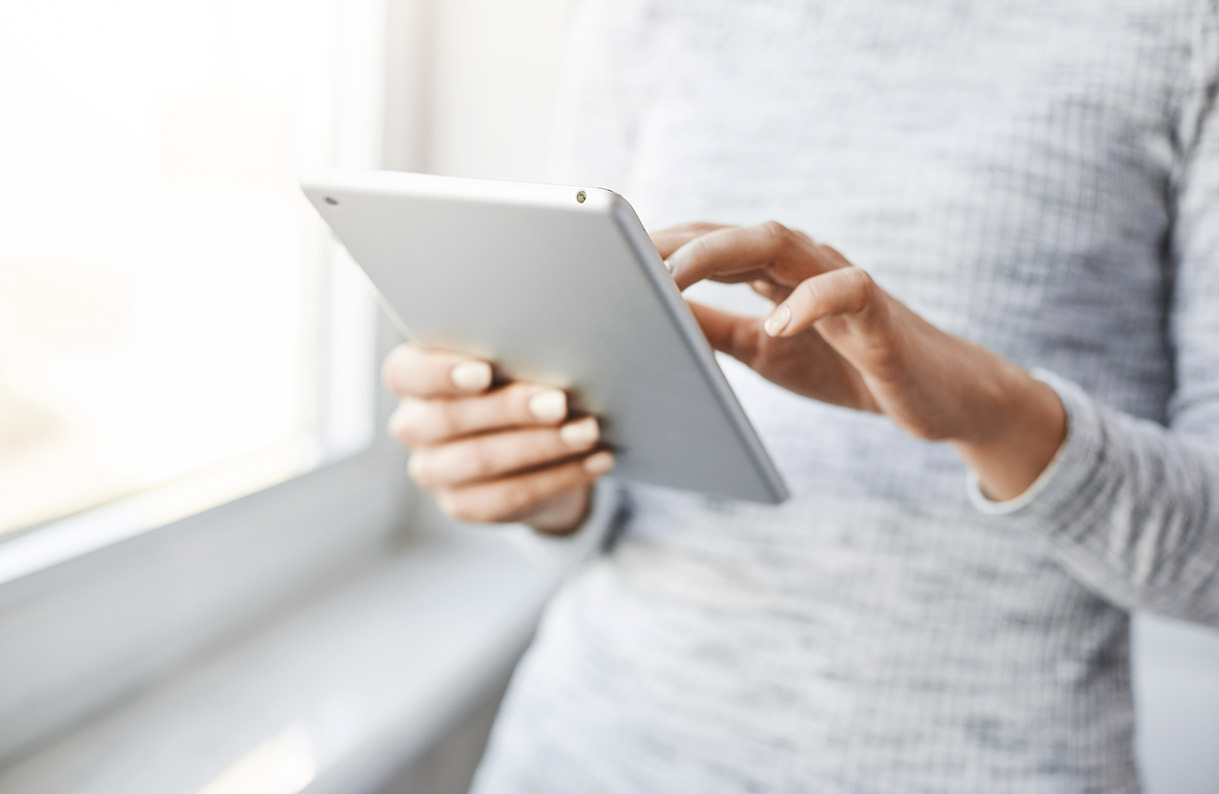 Una persona usa una tableta plateada frente a una ventana luminosa. Lleva una camisa de manga larga gris jaspeada.