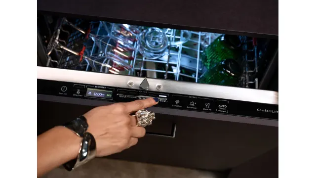 Una mano presiona el botón de una pantalla táctil de un elegante lavavajillas, revelando una rejilla superior abierta con platos dentro.