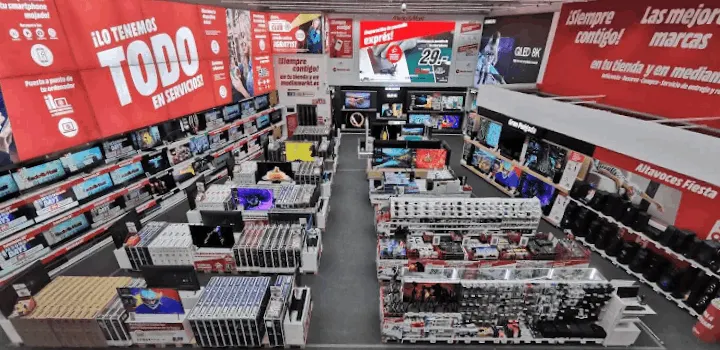 Interior de tienda con estantes llenos de televisores, altavoces y otros productos electrónicos. Hay carteles en rojo y blanco.