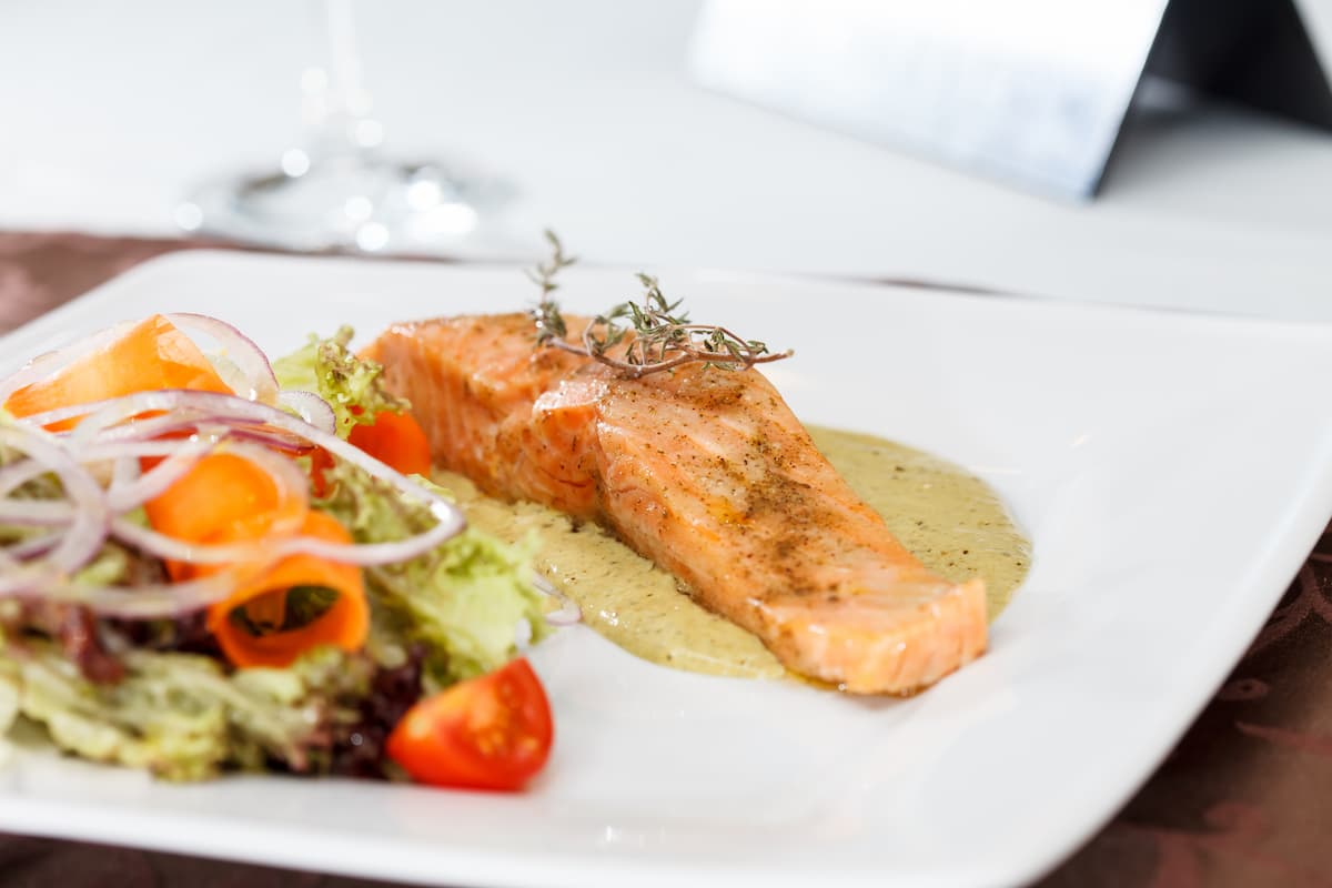 Filete de salmón horneado con salsa verde y ensalada fresca servido en un plato blanco de restaurante.