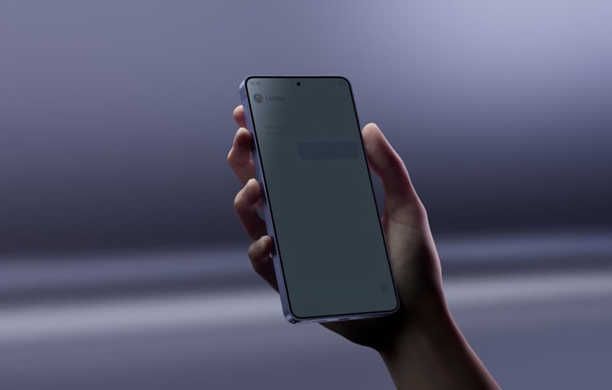 Mano sosteniendo un Samsung Galaxy S26 con la pantalla encendida sobre un fondo desenfocado y gris.