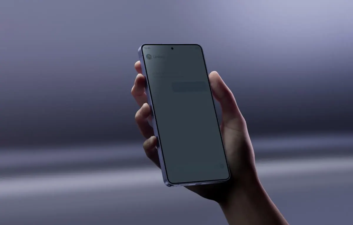 Mano sosteniendo un Samsung Galaxy S26 con la pantalla encendida sobre un fondo desenfocado y gris.