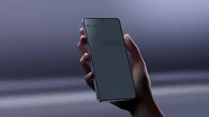 Mano sosteniendo un Samsung Galaxy S26 con la pantalla encendida sobre un fondo desenfocado y gris.