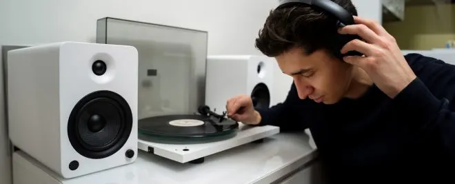 Chico con auriculares de diadema mientras coloca la aguja sobre un disco de vinilo en un tocadiscos, acompañado de dos altavoces blancos