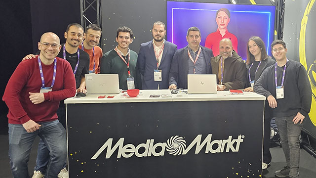 Nueve personas posan tras un mostrador negro con el logotipo de MediaMarkt en blanco. Hay portátiles y una pantalla iluminada detrás.