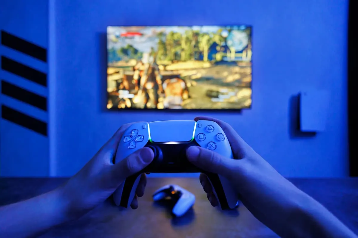 Manos sosteniendo un mando de videojuegos frente a un televisor con un juego en pantalla y luz azul ambiente.