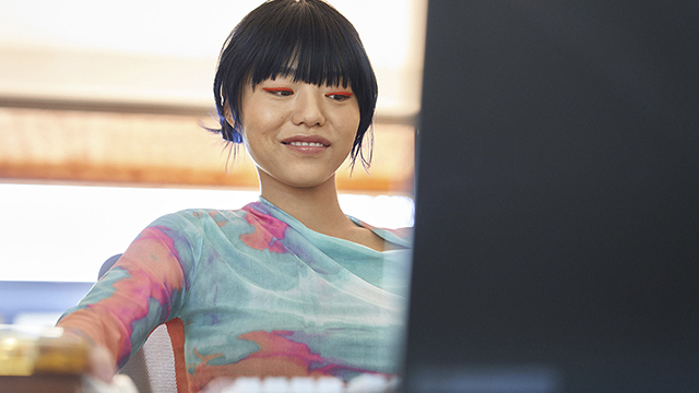 Una persona con flequillo mira una computadora portátil. La persona lleva una blusa azul, rosa y naranja. Hay persianas enrollables en el fondo.