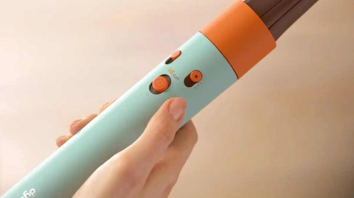 Mano sujetando rizador de pelo turquesa y naranja con botones. El aparato tiene un extremo marrón. Fondo beige.