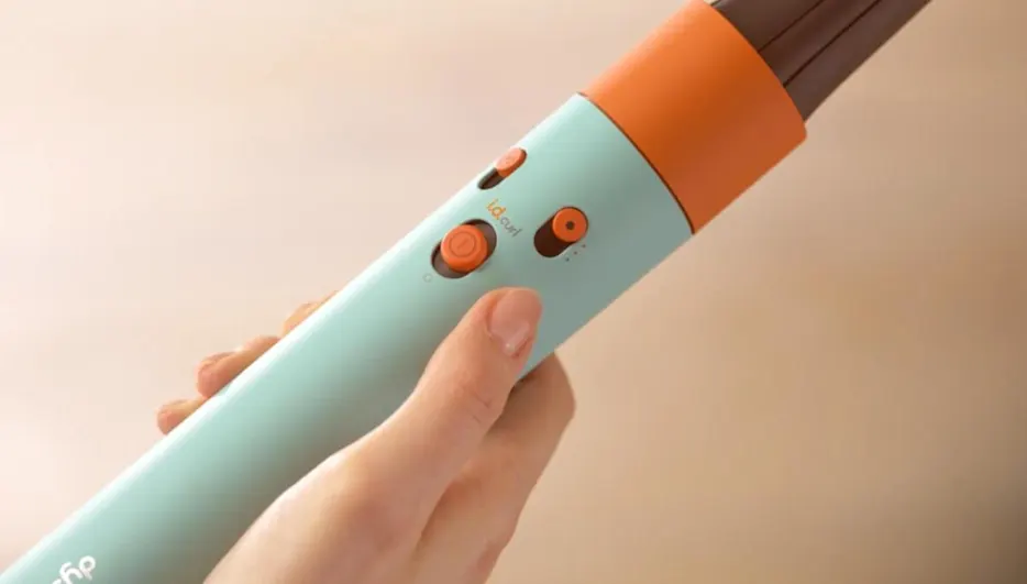 Mano sujetando rizador de pelo turquesa y naranja con botones. El aparato tiene un extremo marrón. Fondo beige.