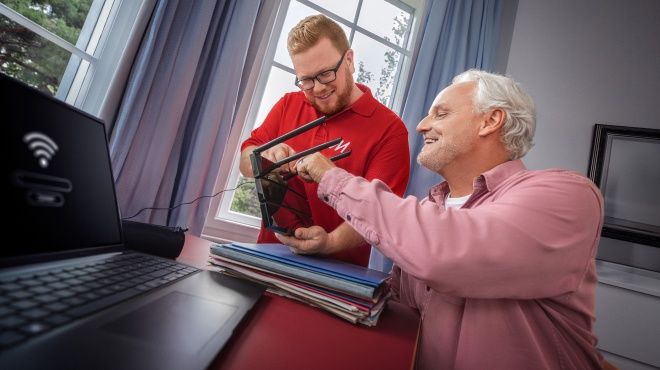 Vendedora explica un producto rojo a una pareja sentada en un sofá. Hay una planta en maceta y una manta roja en el fondo.
